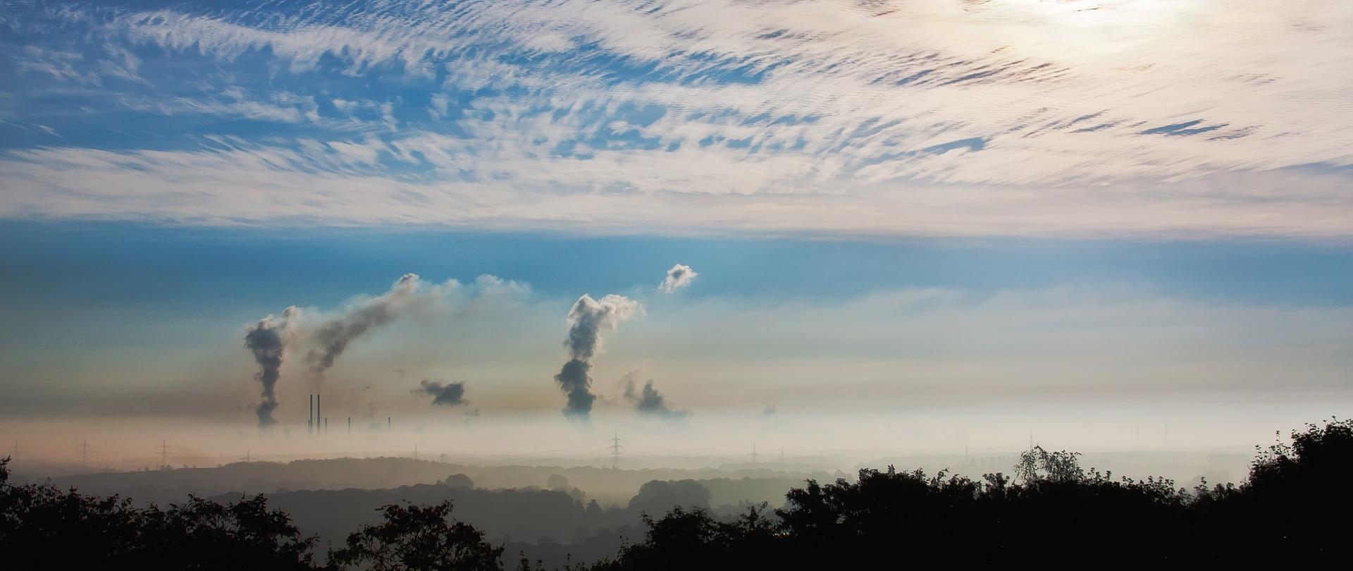 Niebieskie niebo na którym znajdują się białe chmury. W tle widać dymiące kominy.