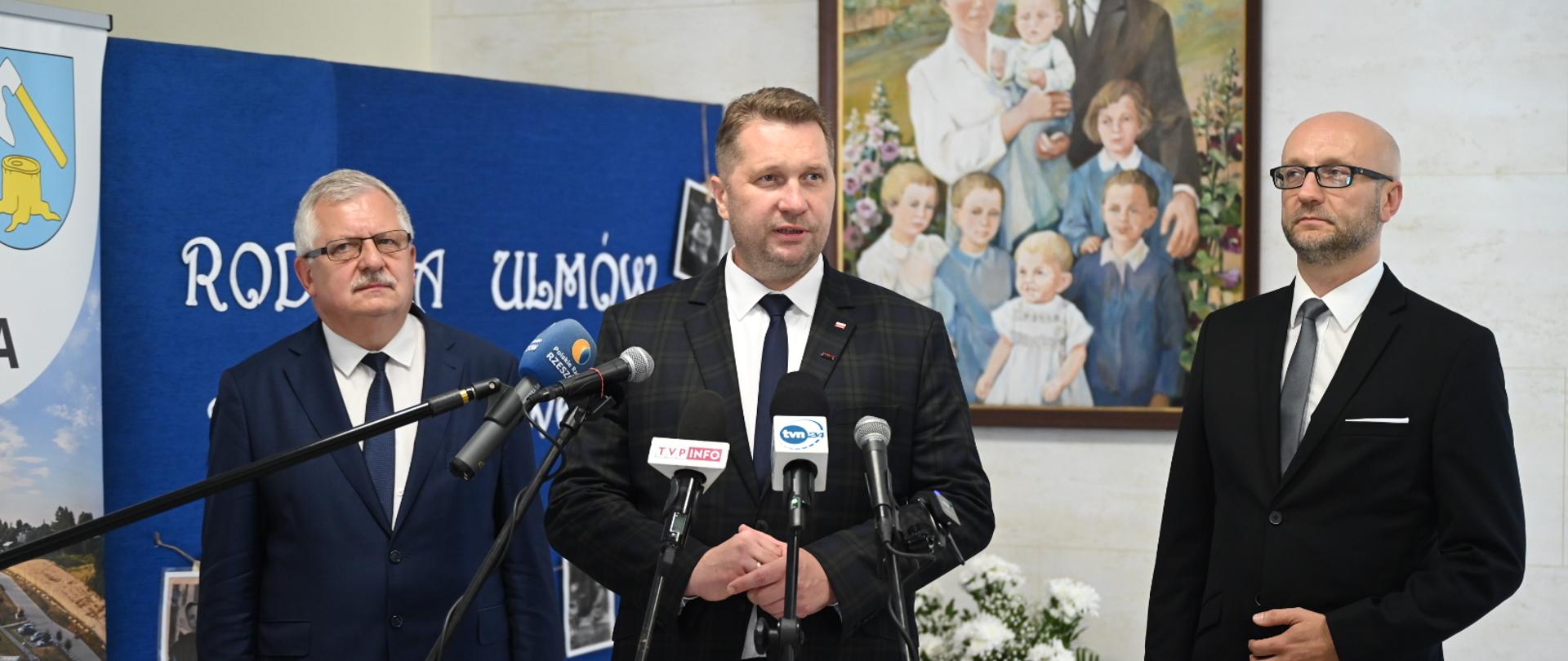 Minister Czarnek stoi pod ścianą na której wisi rodzinny portret, obok niego dwóch mężczyzn w garniturach, przed nimi kilka mikrofonów.