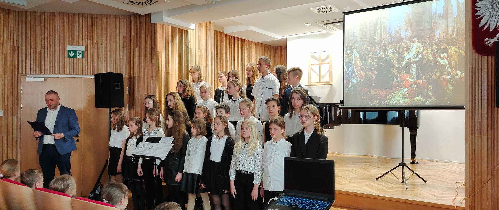 Zdjęcie przedstawia stojących w czasie występu na scenie szkoły naucz. Karola Palicę i chór szkolny