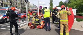 Na zdjęciu widać strażaków ubranych w piaskowe oraz czarne ubrania ochronne zabezpieczonych w maseczki chirurgiczne , którzy udzielają kwalifikowanej pomocy medycznej osobie poszkodowanej. Na chodniku leżą czerwono-żółte torby PSP R-1 .Na zdjęciu znajduje się też Policjant ubrany w żółtą kamizelkę. Na drugim planie znajdują się dwa czerwone samochody PSP oraz biało-czerwono-niebieska karetka Pogotowia ratunkowego. 