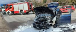 Samochód osobowy z podniesioną maską i pokryty pianą gaśniczą. W tle samochód pożarniczy oraz jadące samochody osobowy i ciężarowy.