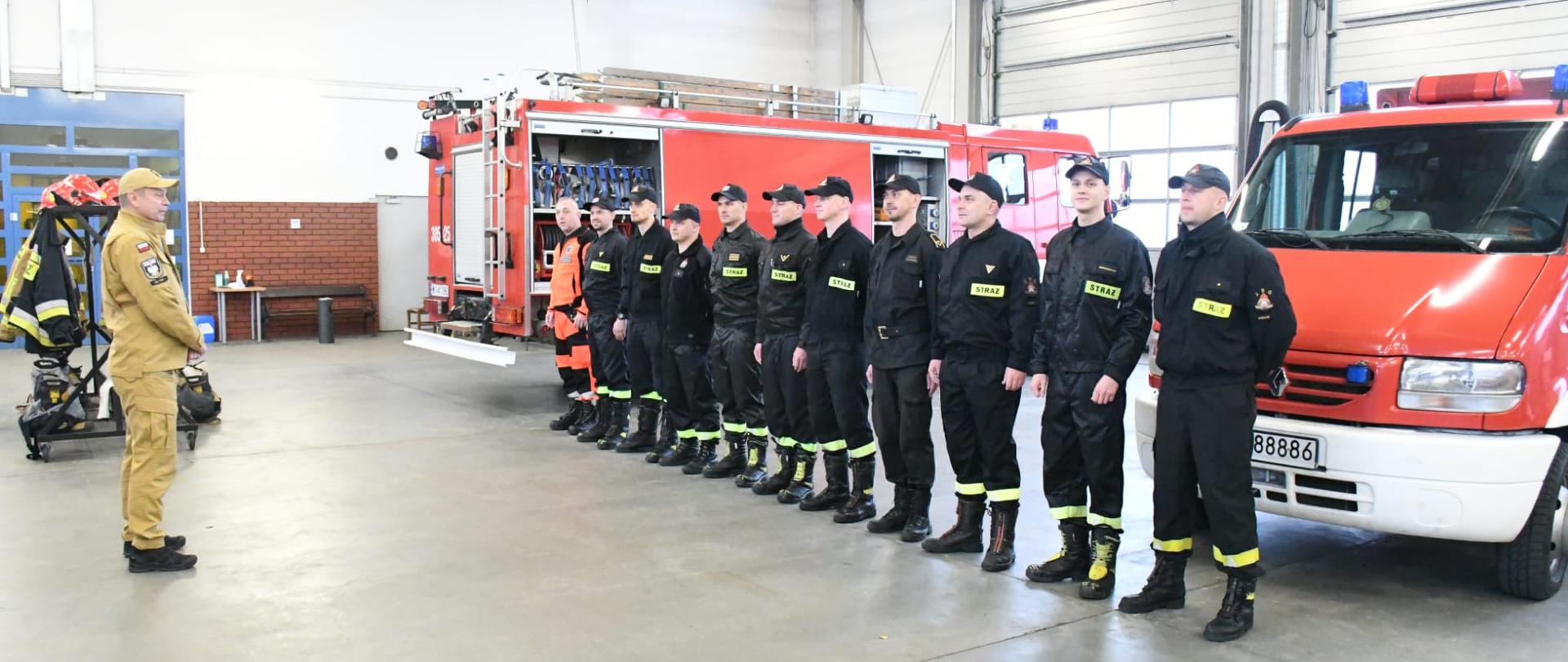 Po lewo stoi Zastępca KG PSP który przeprowadza odprawę z ratownikami medycznymi PSP którzy stoją po prawo w szeregu, w tle samochody PSP, wszyscy znajdują się w garażu jednostki ratowniczo - gaśniczej