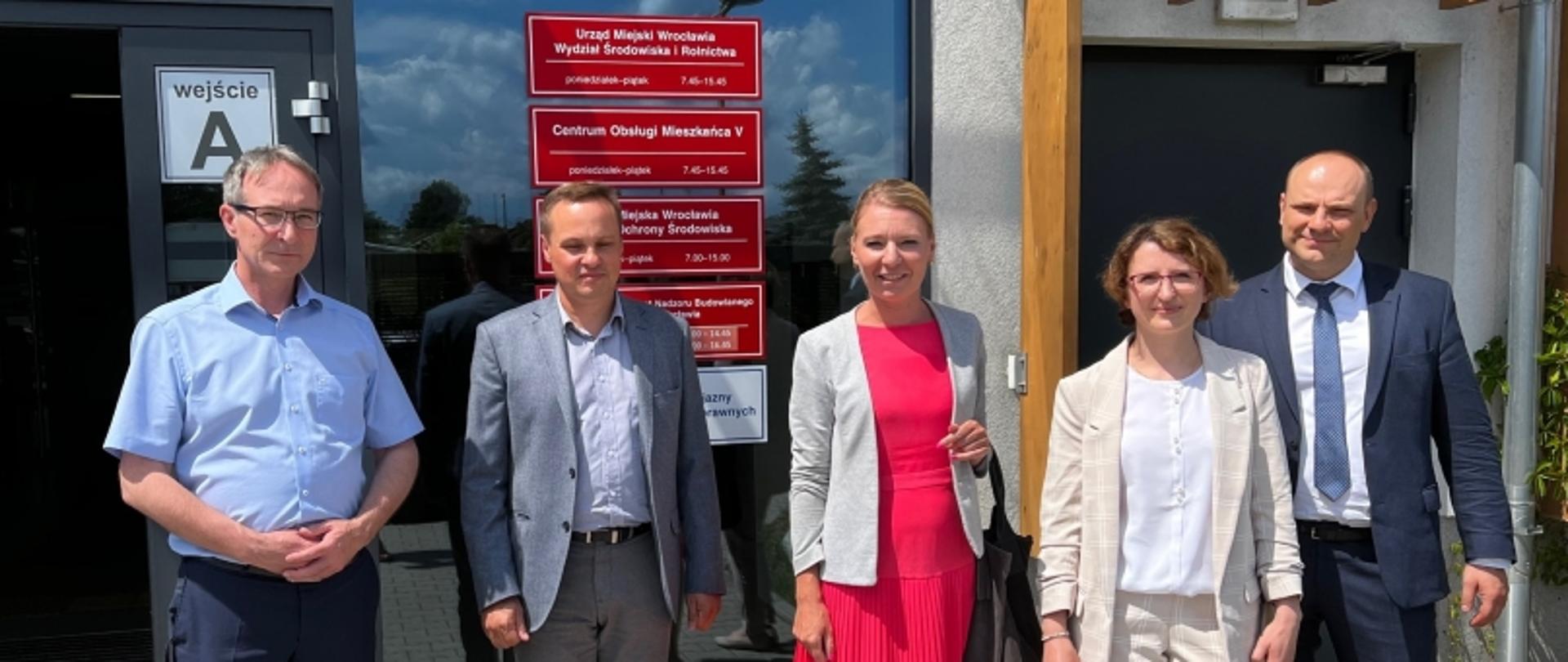 Pięć osób stojących przed wejściem „A” do budynku Urzędu Miejskiego Wrocławia. Na drzwiach widoczne są czerwone tabliczki informujące o godzinach pracy i lokalizacji różnych wydziałów, w tym Wydziału Środowiska i Rolnictwa oraz Zarządu Zieleni Miejskiej.