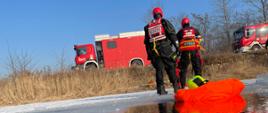 Na pierwszym planie przerębel, dalej dwóch ratowników. W tle widoczne wozy strażackie.