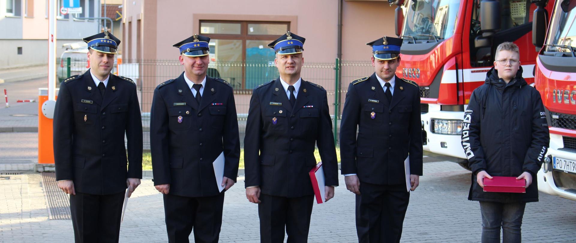 Zdjęcie przedstawia strażaków podczas uroczystej zmiany służby.
W tle samochody strażackie i budynek JRG.
