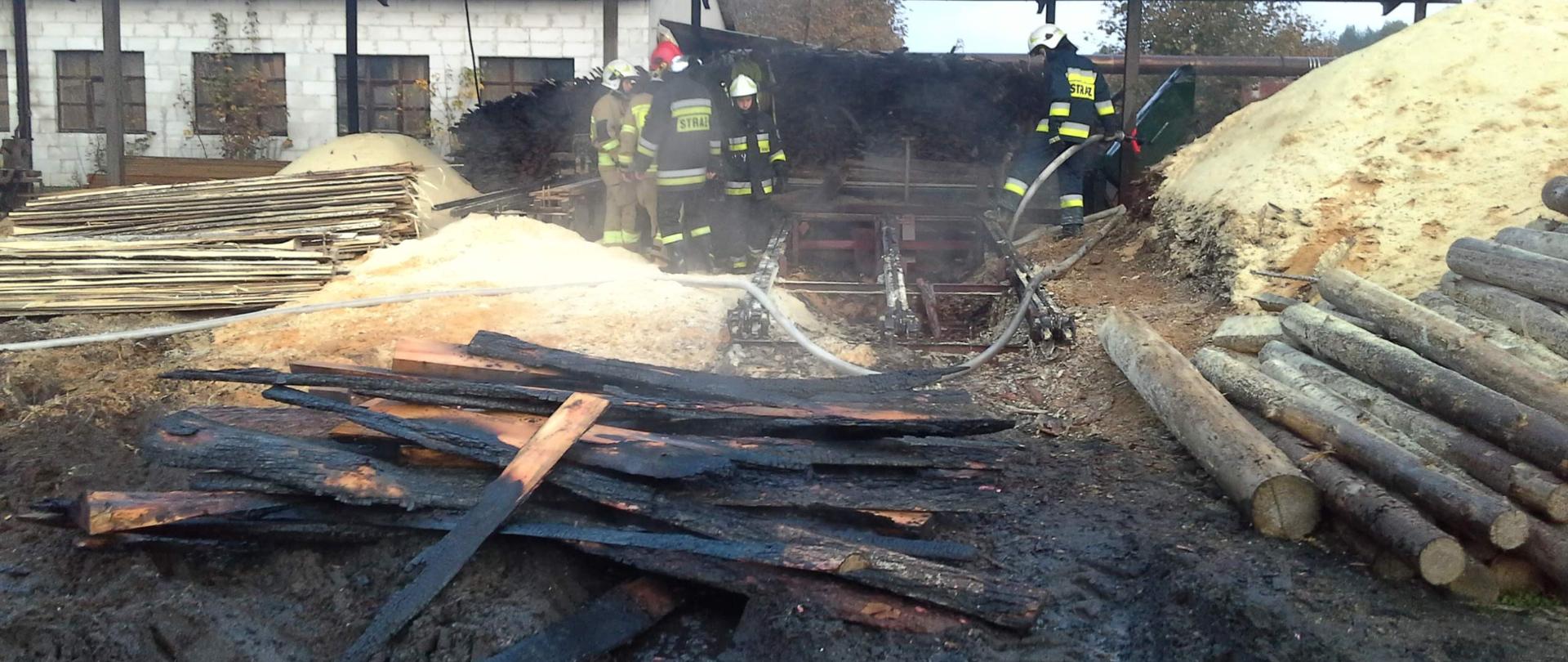Na pierwszym planie spalone deski leżące w błocie, w oddali sześcioro pracujących strażaków, wokoło widać składowane deski jak i hałdy trocin. W tle zadaszenie tartaku.