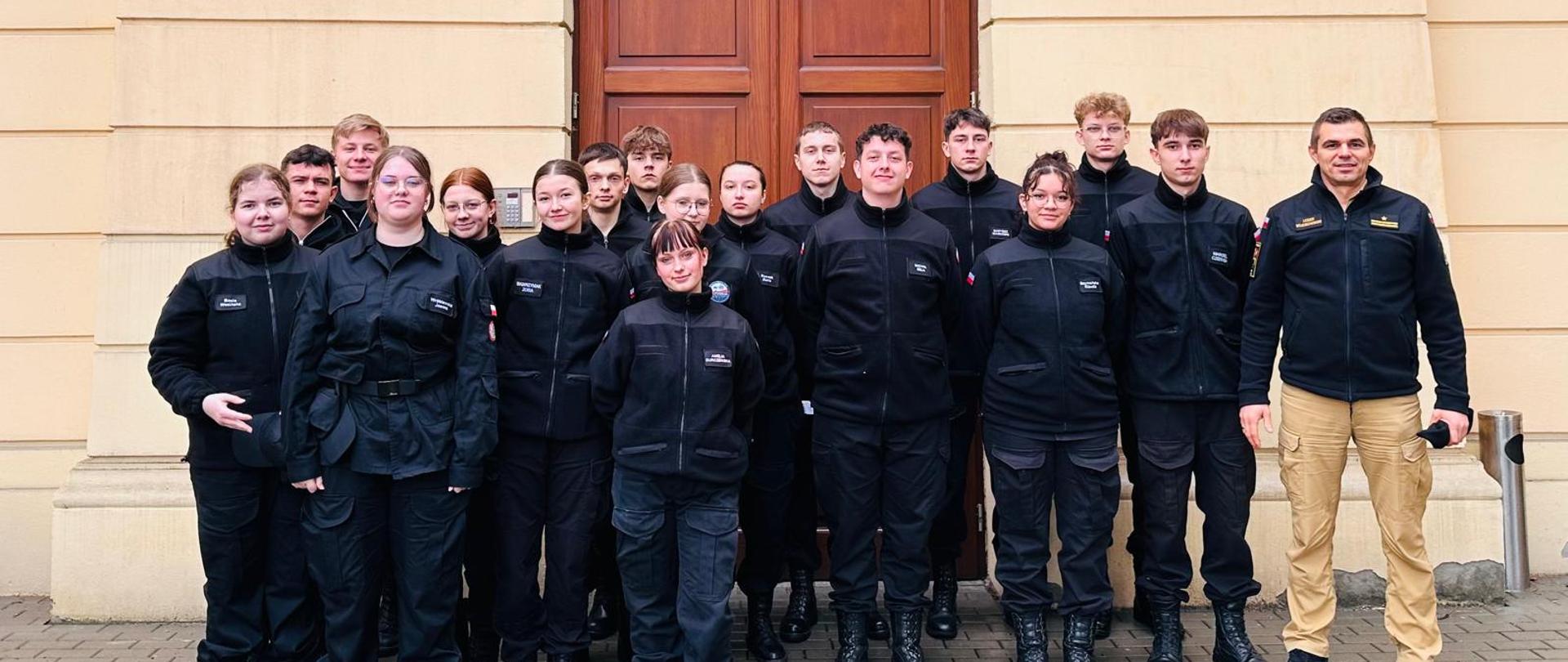 Na zdjęciu widać grupę osób wraz z funkcjonariuszem przed Centrum Edukacji i Historii Warszawskiej Straży Pożarnej