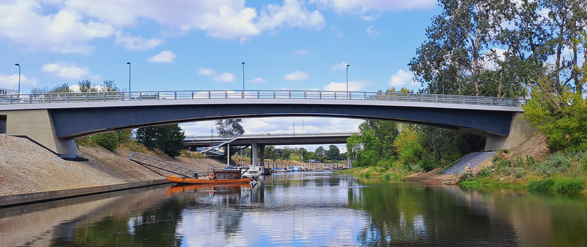 Na zdjęciu widać betonową kładkę nad kanałkiem czerniakowskim. W oddali na wodzie widać łódki. Na prawym brzegu widoczne zarośla i drzewa. 