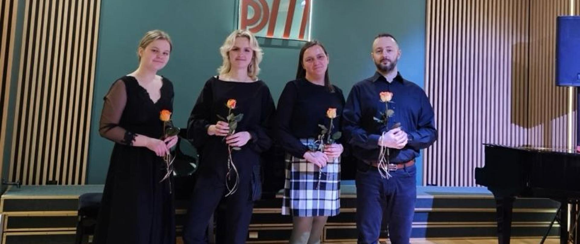 Na zdjęciu widoczne są cztery osoby stojące na scenie w eleganckiej sali koncertowej. Każda z nich trzyma pojedynczą pomarańczową różę z ozdobną wstążką, co sugeruje, że jest to moment podziękowań po występie.
Osoby są ubrane w eleganckie, ciemne stroje. Pierwsza kobieta po lewej stronie ma na sobie długą, czarną suknię z przezroczystymi rękawami. Druga kobieta, stojąca obok, ubrana jest w czarny strój i botki na obcasie. Trzecia kobieta nosi czarny sweter i spódnicę w kratę, a także zielone rajstopy i czarne buty. Ostatnia osoba, mężczyzna, ubrany jest w ciemną koszulę, czarne spodnie i brązowe buty.
W tle znajduje się logo z literami „PSM”, co wskazuje na szkołę muzyczną. Obok nich widoczny jest fortepian, a całe wnętrze ma nowoczesny, drewniany wystrój.