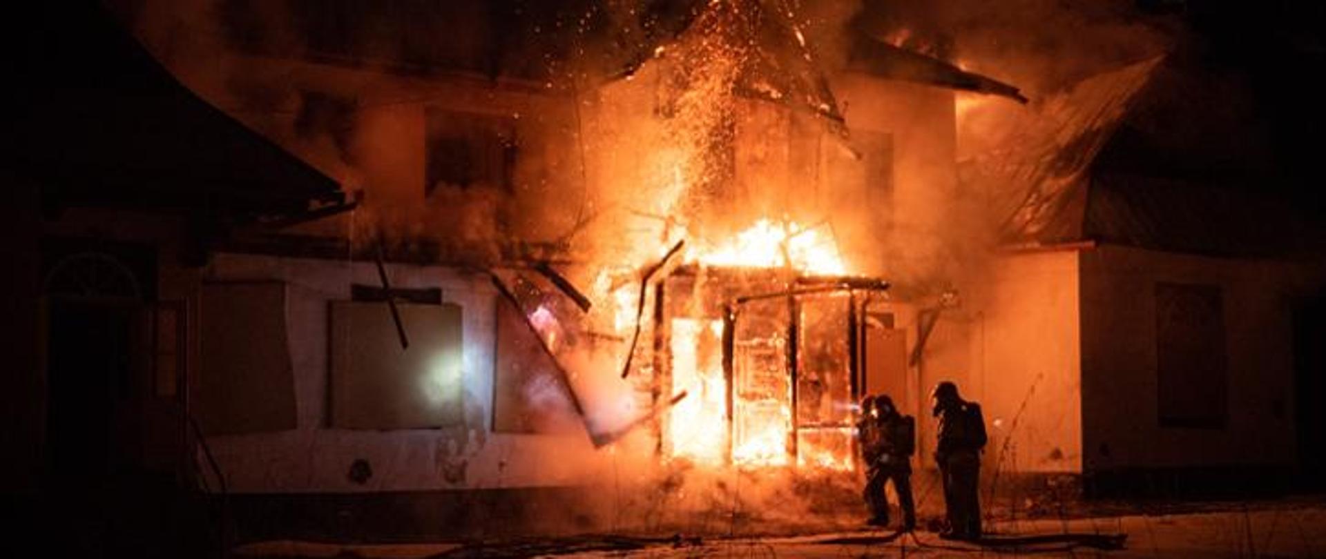 Noc. Budynek mieszkalny stojący w płomieniach. Przed nim dwóch strażaków.