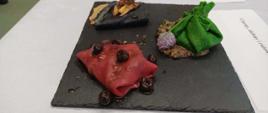 Zdjęcie przedstawia elegancko zaserwowane danie restauracyjne, składające się z trzech różnokolorowych naleśników, podanych na czarnej, kwadratowej płycie łupkowej o nieregularnych krawędziach. Płyta leży na białym obrusie.Główne elementy potrawyNaleśnik różowy (na pierwszym planie po lewej): ma intensywny, bordowo-różowy kolor (prawdopodobnie z dodatkiem soku z buraka). Jest złożony w trójkąt, polany błyszczącym sosem i udekorowany kilkoma ciemnymi, okrągłymi owocami (wyglądają jak wiśnie lub borówki).Naleśnik zielony (po prawej stronie): ma jaskrawozielony kolor (prawdopodobnie z dodatkiem szpinaku). Został uformowany w sakiewkę, związaną na górze zielonym szczypiorkiem. Sakiewka stoi na ciemniejszym farszu (wyglądającym na grzybowy lub mięsny), a obok niej leży jadalny, fioletowy kwiat szczypiorku.Naleśnik czarny (w tle po lewej): ma bardzo ciemny, niemal czarny kolor (prawdopodobnie z dodatkiem sepii lub aktywnego węgla). Jest zwinięty w rulon lub złożony w prostokąt, podany na jasnobrązowym sosie i posypany z góry podsmażoną cebulką lub kawałkami warzyw.Tło i kontekstPo prawej stronie, na białym obrusie, widoczny jest fragment białej kartki z wydrukowanym napisem w języku polskim. Można odczytać fragment tekstu: „Czarny, zielony i różowy”.Danie wygląda na potrawę konkursową lub pokazową, przygotowaną z dbałością o estetykę i kontrast kolorów.