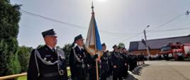 Kolorowa fotografia wykonana w słoneczny dzień przedstawia szerokie ujęcie pododdziału strażaków wraz z pocztem sztandarowym, ubranych na galowo stojących na placu przed remizą strażacką podczas uroczystej zbiórki. W tle widać część czerwonego wozu bojowego straży pożarnej i zabudowania przy których stoją osoby cywilne.
