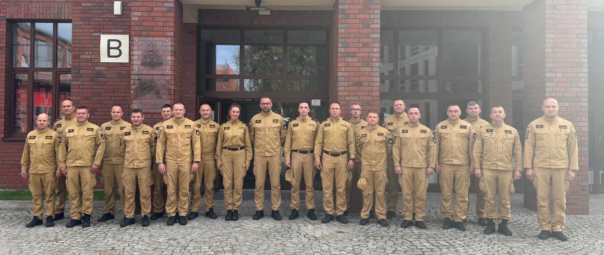 Grupa funkcjonariuszy Państwowej Straży Pożarnej w jasnobrązowych mundurach służbowych pozuje do pamiątkowego zdjęcia na tle ceglanego budynku Komendy Wojewódzkiej Państwowej Straży Pożarnej. Strażnicy stoją w jednym rzędzie na tle wejścia i elewacji z widocznym napisem „KOMENDA WOJEWÓDZKA PAŃSTWOWEJ STR...”. Wśród uczestników widoczni są zarówno mężczyźni, jak i kobieta, wszyscy w jednakowych uniformach z czarnym obuwiem.