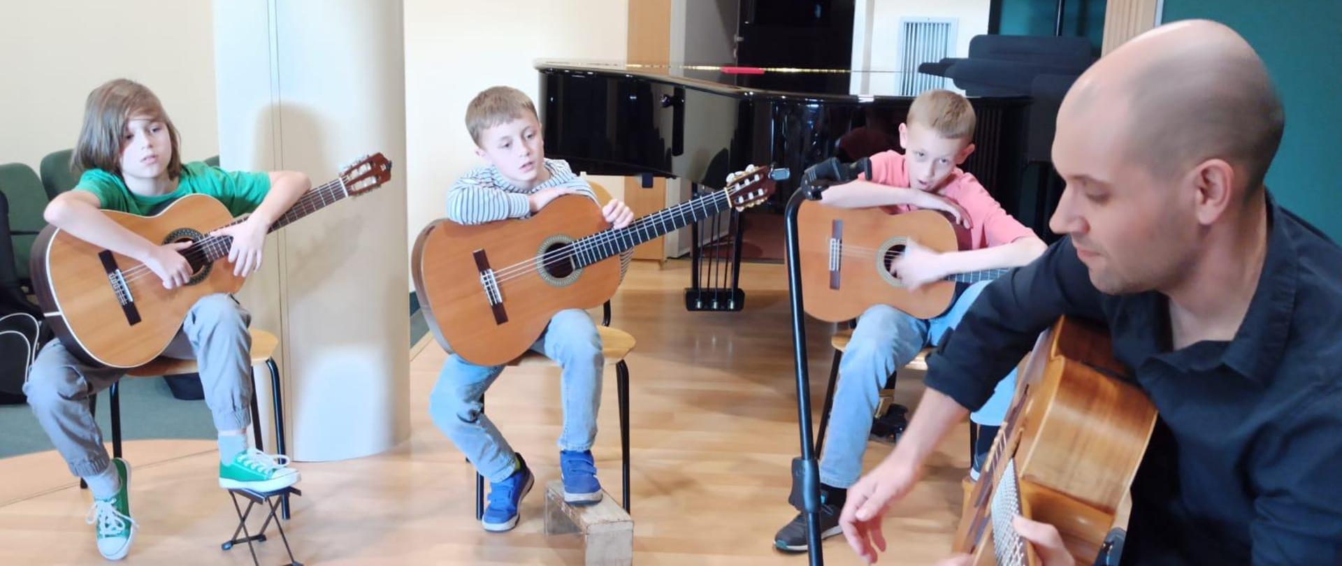 Na zdjęciu widać uczniów klasy gitary oraz Piotra Krępecia który prowadził warsztaty. Uczniowie trzymają gitary a nauczyciel gra na gitarze. W tle widać fortepian po lewej stronie parę siedzeń z publiczności w tle żółta ściana a po prawej stronie zielona ściana