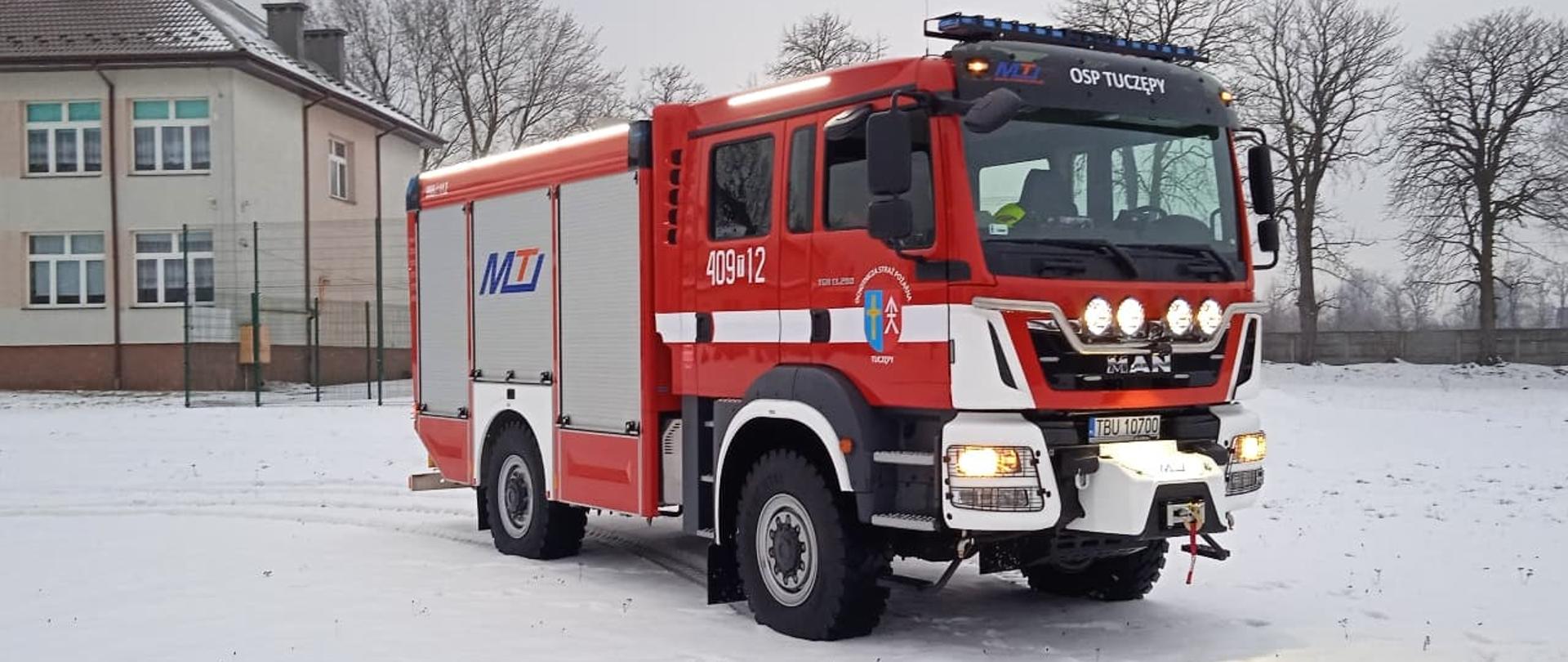 Samochód ratowniczo-gaśniczy marki MAN w kolorze czerwonym, z lewej strony zdjęcia fragment budynku szkoły podstawowej.