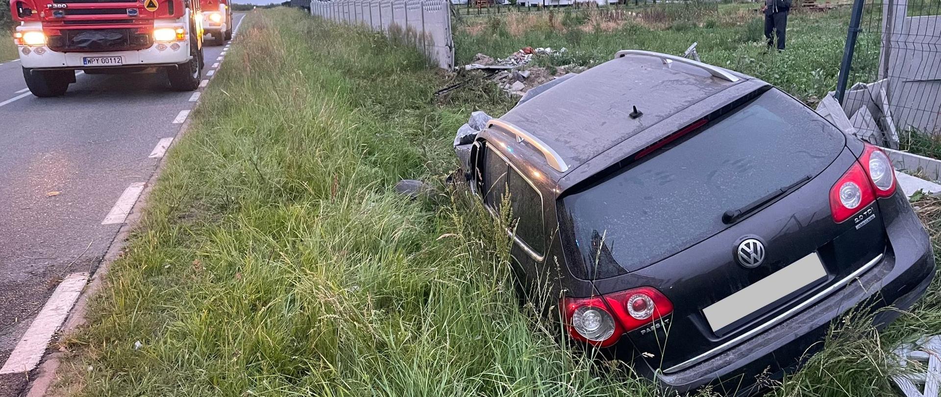 Zdjęcie przedstawia ciemny samochód osobowy, który wypadł z drogi i leży przewrócony na boku w głębokim rowie, porośniętym wysoką trawą. Z tyłu samochodu widoczne jest logo Volkswagena. Na drodze, po lewej stronie, stoi czerwony wóz strażacki marki Scania z włączonymi światłami awaryjnymi. W tle widać wiejskie domy i drzewa. W oddali, po prawej stronie, stoi mężczyzna. Niebo jest pochmurne.