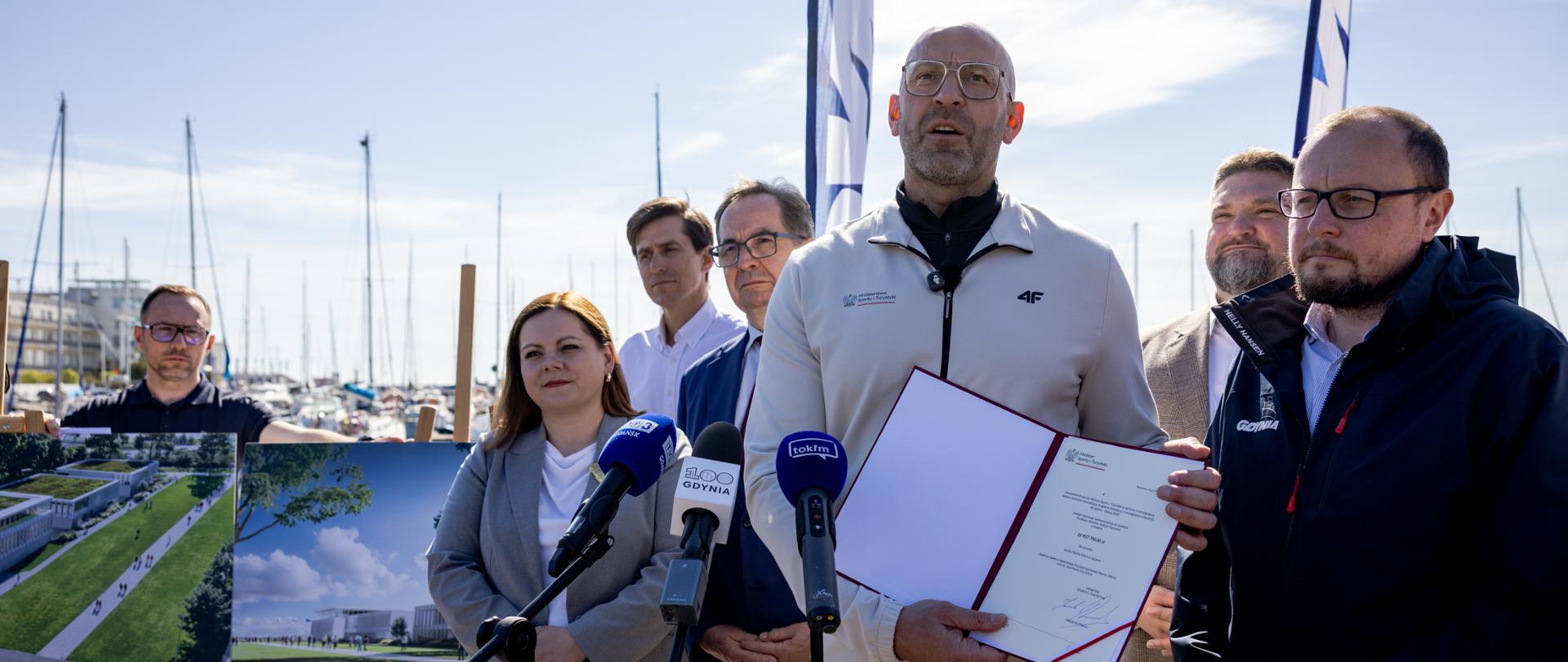 Minister sportu i turystyki Jakub Rutnicki prezentuje decyzję o dofinansowaniu budowy Olimpijskiego Centrum Żeglarstwa w Gdynie. Zdjęcie zrobione podczas konferencji prasowej na świeżym powietrzu. Z tyłu stoją inni uczestnicy konferencji. Z lewej strony z tyłu widać plansze z wizualizacjami inwestycji. W tle widać maszty żaglówek.
