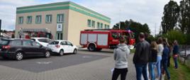 Grupa ewakuowanych osób stoi ustawiona po prawej stronie budynku. Przed budynkiem stoją dwa samochody ratowniczo-gaśnicze. Na parkingu stoją zaparkowane pojazdy.