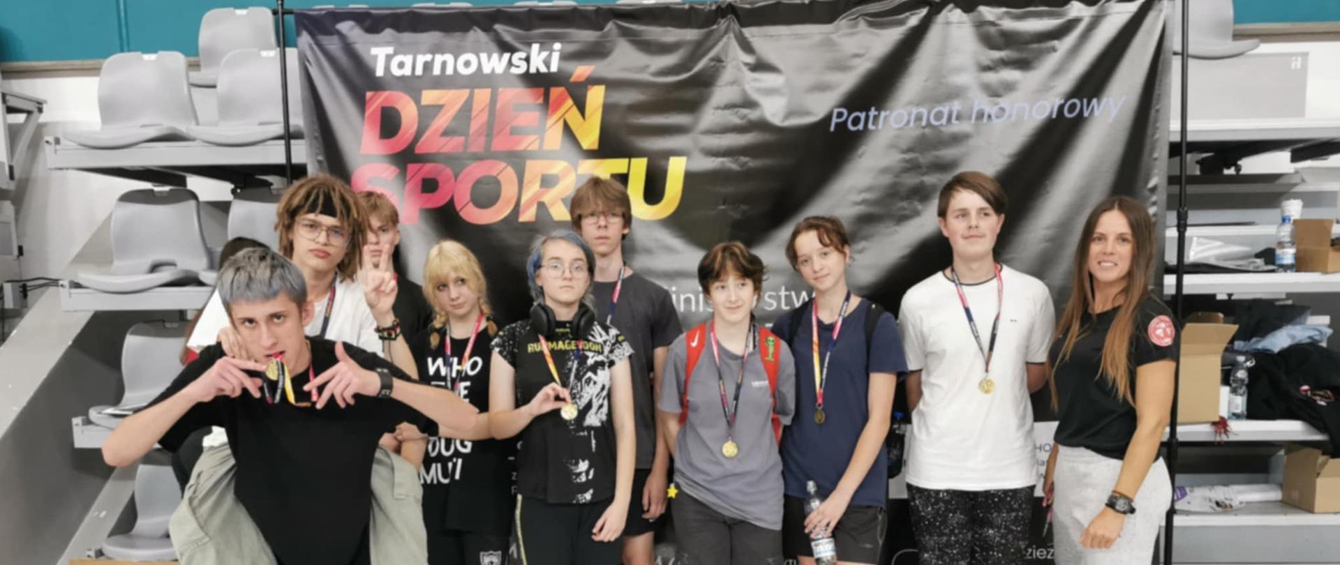 Grupa młodzieży z medalami sportowymi pozuje na tle baneru Tarnowski Dzień Sportu, obok stoi trenerka.