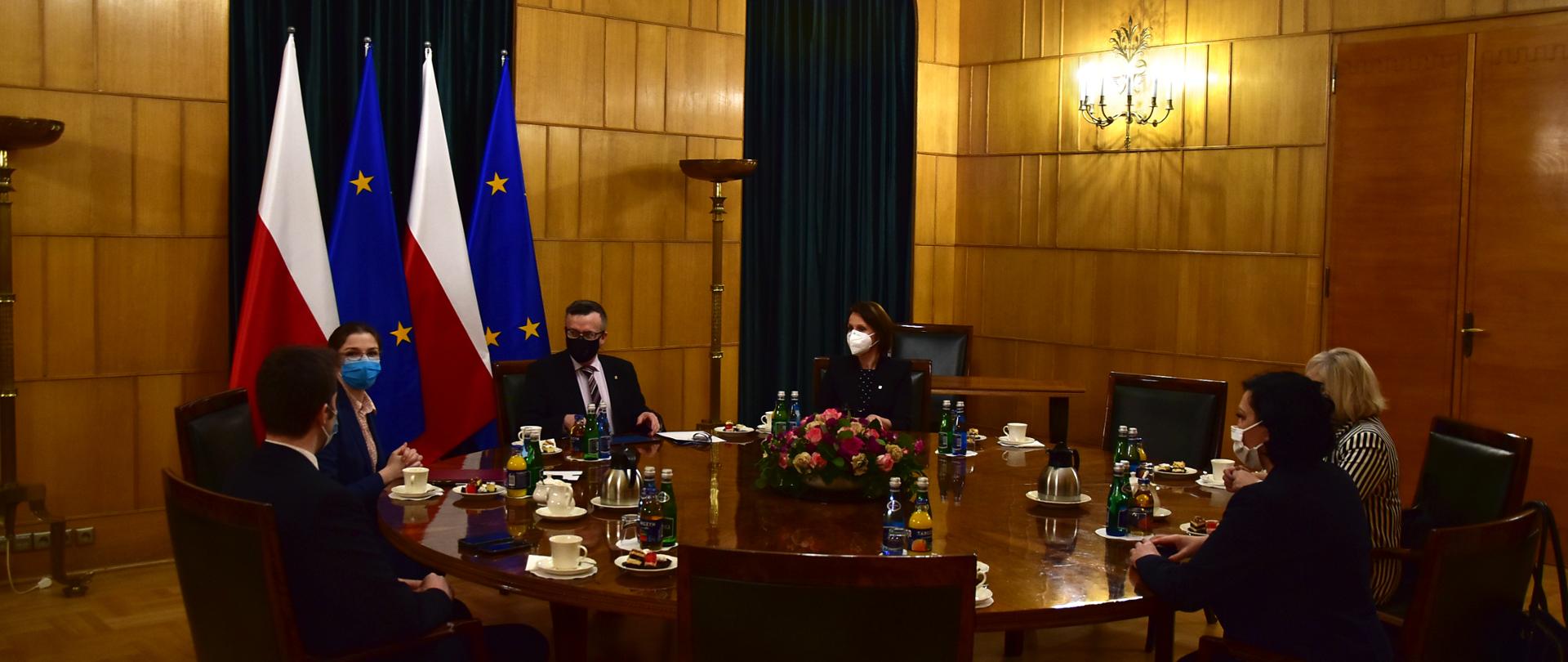 Uczestnicy spotkania siedzą przy okrągłym stole. Za Szefem Służby Cywilnej flagi: Polski i Unii Europejskiej