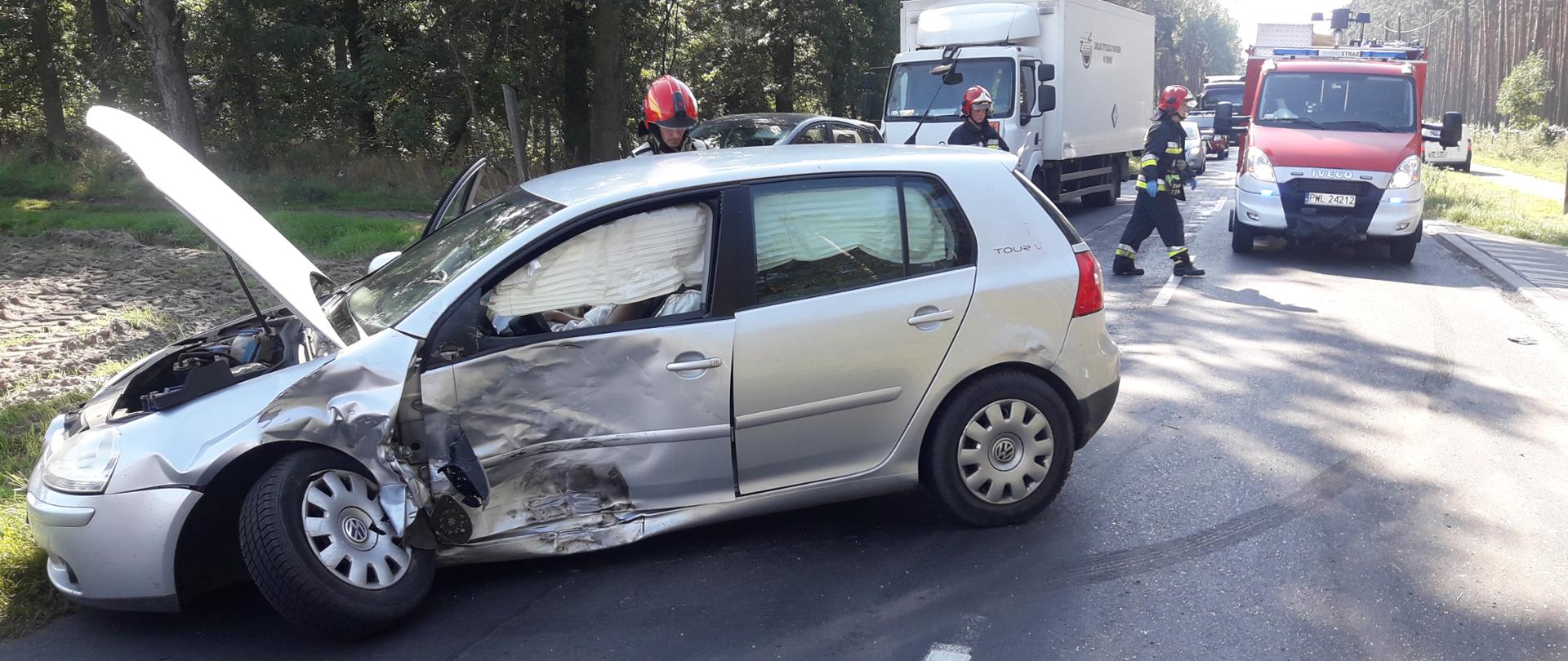 Jeden z samochodów biorący udział w kolizji stoi rozbity na pasie drogowym blokując ruch. W tle strażacy, zabezpieczający miejsce zdarzenia.