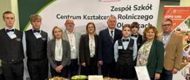 Zdjęcie przedstawia grupę dziewięciu osób – uczniów oraz nauczycieli i przedstawicieli Zespołu Szkół Centrum Kształcenia Rolniczego w Oleszycach. Pomiędzy nimi stoi dyrekcja i kadra pedagogiczna oraz goście w eleganckich strojach galowych, a po obu stronach – uczniowie w strojach kelnerskich (czarnych kamizelkach i muszkach). 