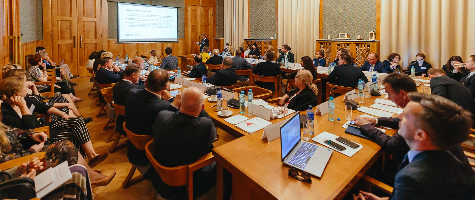 W sali ze ścianami wyłożonymi drewnianymi panelami przy ustawionych w czworokąt stołach siedzi dużo ludzi, w z tyłu na ścianie duży ekran.