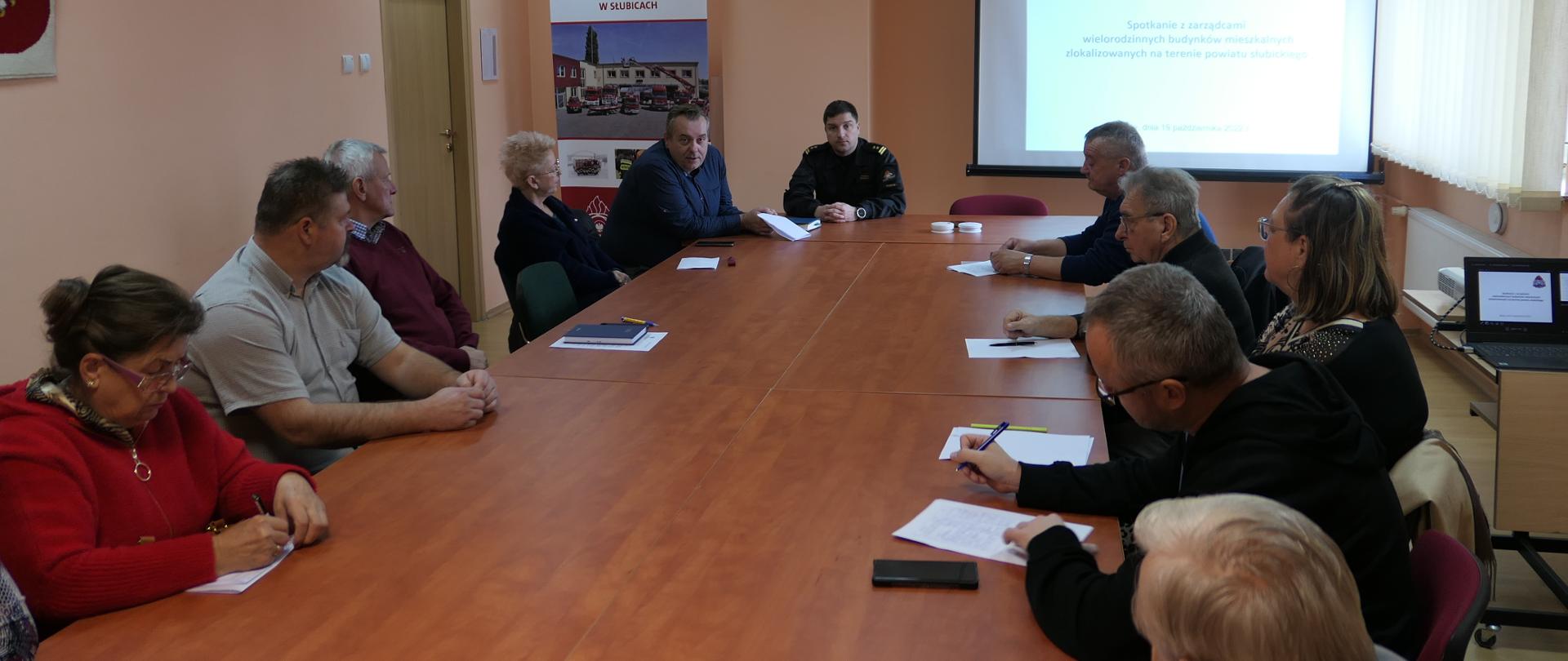 Wnętrz pomieszczenia świetlicy w KP PSP Słubice. Na środku duży stół wokół którego siedzą osoby. U góry umundurowany funkcjonariusz PSP a obok niego baner KP PSP w Słubicach oraz ekran na którym wyświetlana jest prezentacja.