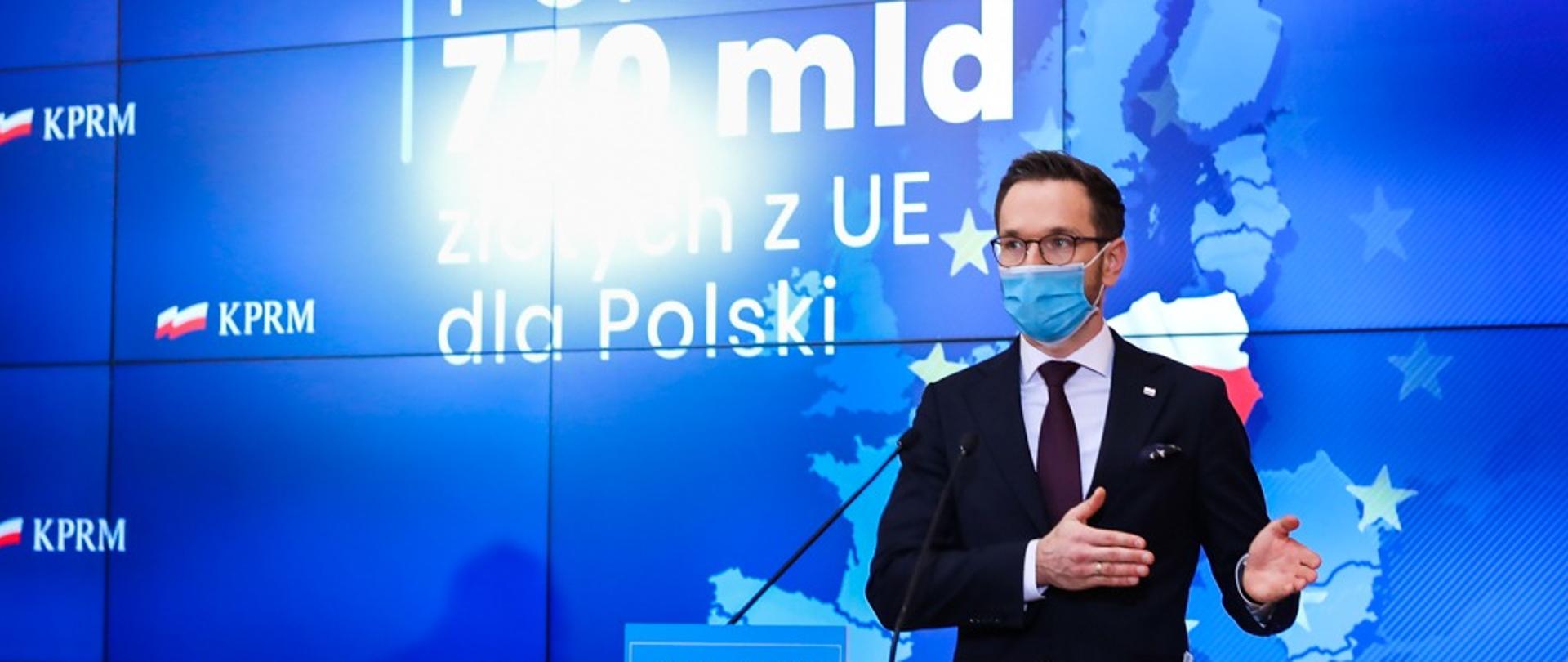 Wiceminister Waldemar Buda stoi w maseczce przed mównicą. Za plecami ekran z napisem: "Ponad 770 mld złotych z UE dla Polski". 