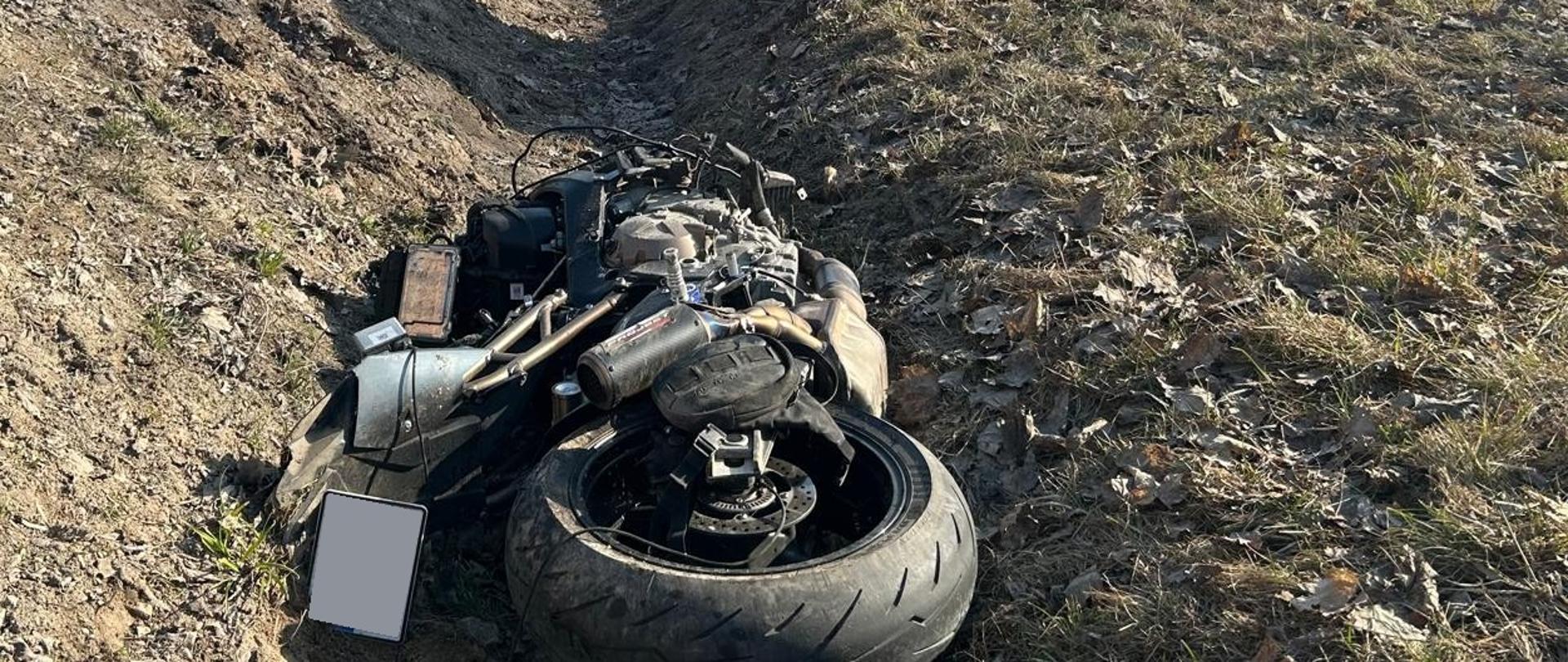 Na pierwszym planie zdjęcia widoczny jest uszkodzony motocykl należący do ofiary oraz parawan osłaniający miejsce, w którym znajduje się poszkodowany. W tle znajdują się strażacy oraz samochód ratowniczo-gaśniczy zabezpieczający miejsce zdarzenia.