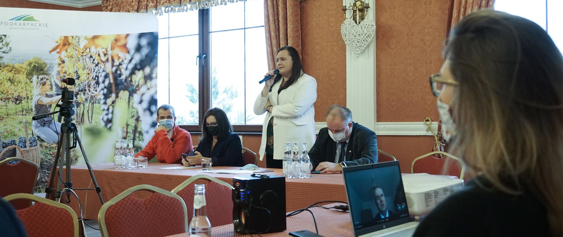 Sala konferencyjna, za stołem siedzą 4 osoby, wiceminister stoi i przemawia do mikrofonu