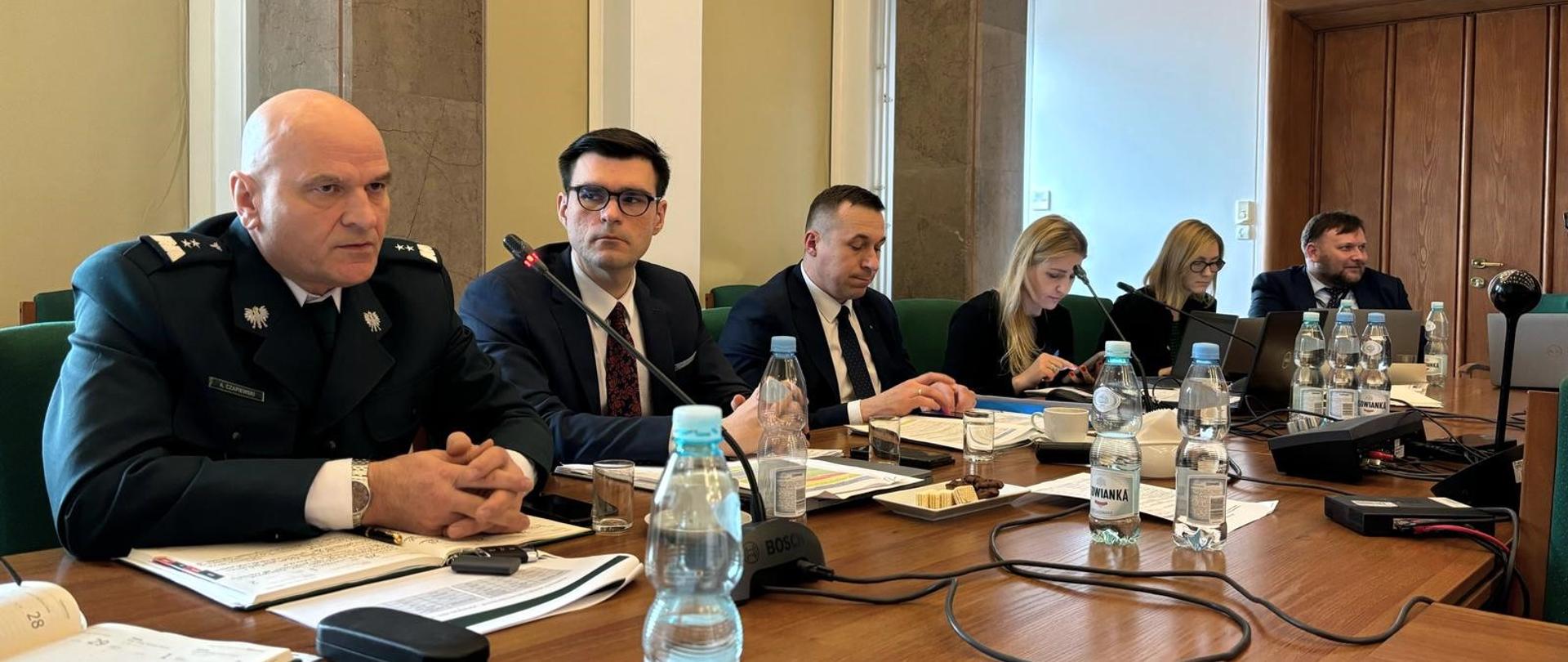 Mężczyzna w mundurze a obok niego za stołem prezydialnym dwóch innych mężczyzn w garniturach i dwie kobiety. 