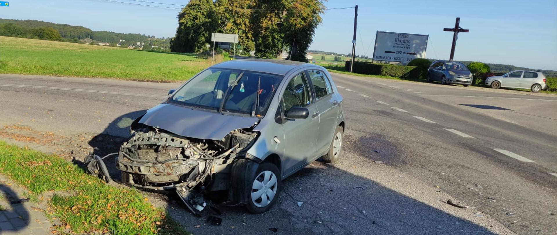 Zdjęcie zrobione w ciągu dnia, słonecznie. Na zdjęciu widać szary samochód osobowy z rozbitym przodem. W tle samochody postronne osobowe pozostawione na poboczu. 