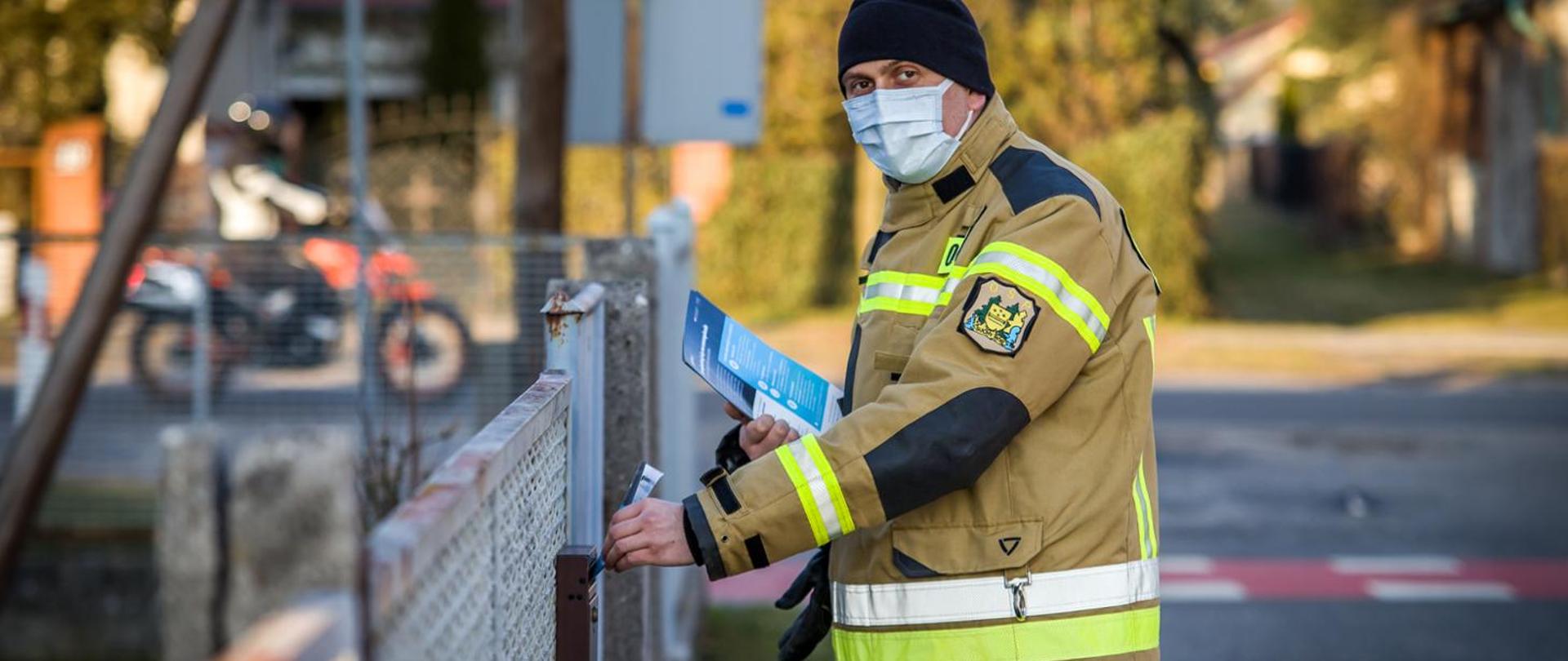 Zdjęcie przedstawia strażaka OSP wrzucającego ulotkę propagujące wiedzę na temat profilaktyki oraz szczepień przeciwko COVID-19. Strażak wrzuca ulotkę lewą ręką do skrzynki na listy. W prawej ręce trzyma pozostałe ulotki. Strażak ubrany w ubranie specjalne koloru piaskowego, czarną czapkę i jasną maseczkę na twarzy.