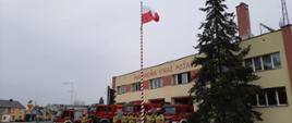 Budynek PSP przed którym na maszt podniesiono Flagę Polską, przed budynkiem wystawiono samochody pożarnicze oraz strażacy ustawieni do uroczystej zbiórki salutują 