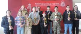 Zdjęcie przedstawia Komendanta Powiatowego PSP w Opatowie ubranego w mundur służbowy koloru żółtego, v-ce Prezesa Zarządu Powiatowego ZOSP RP ubranego w mundur koloru czarnego 9 najlepszych uczestników konkursu z pucharami W tle baner koloru czerwonego z napisem Państwowa Straż Pożarna.