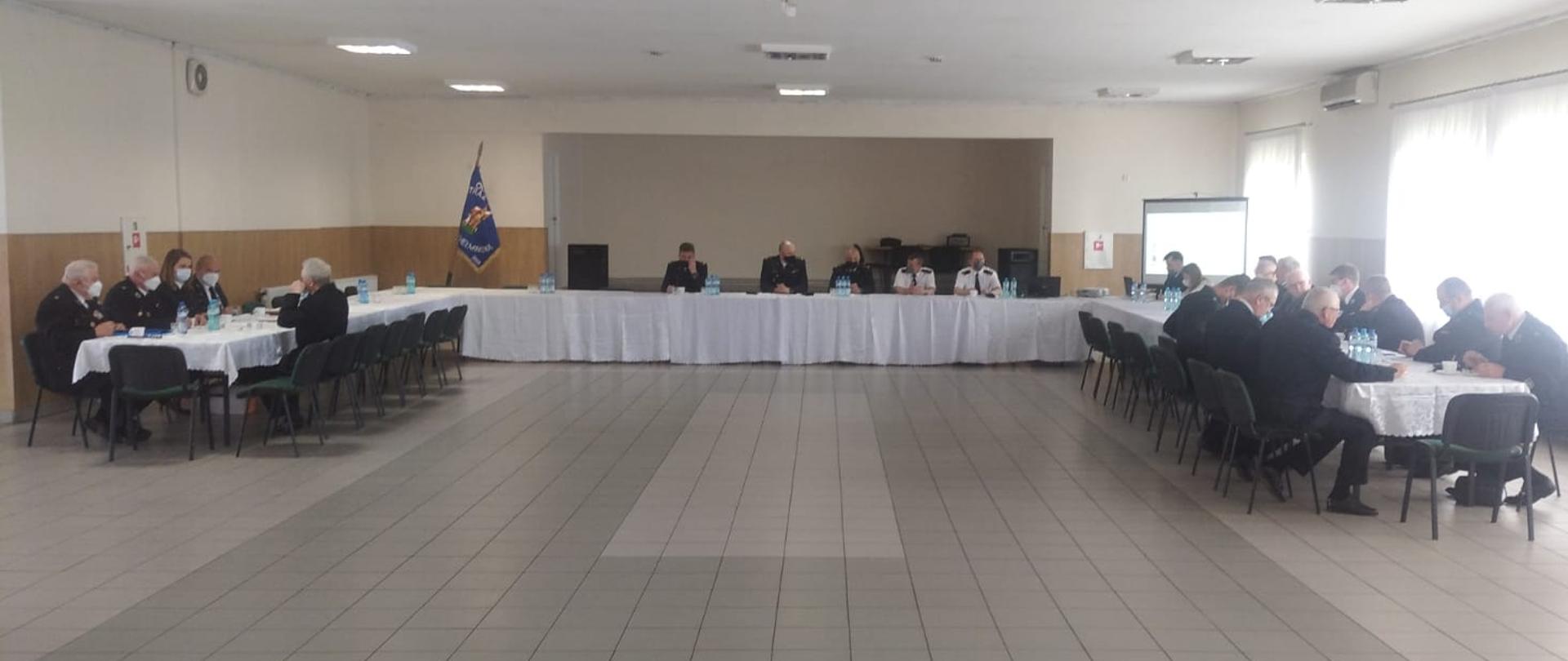 Widok na stoły ustawione w kształcie litery U, przy stołach siedzą przedstawiciele OSP i PSP, wszyscy w swoich mundurach wyjściowych, konsultacje odbywają się w sali OSP Dąbrowa Chełmińska, kremowy kolor ścian, w tle scena.
