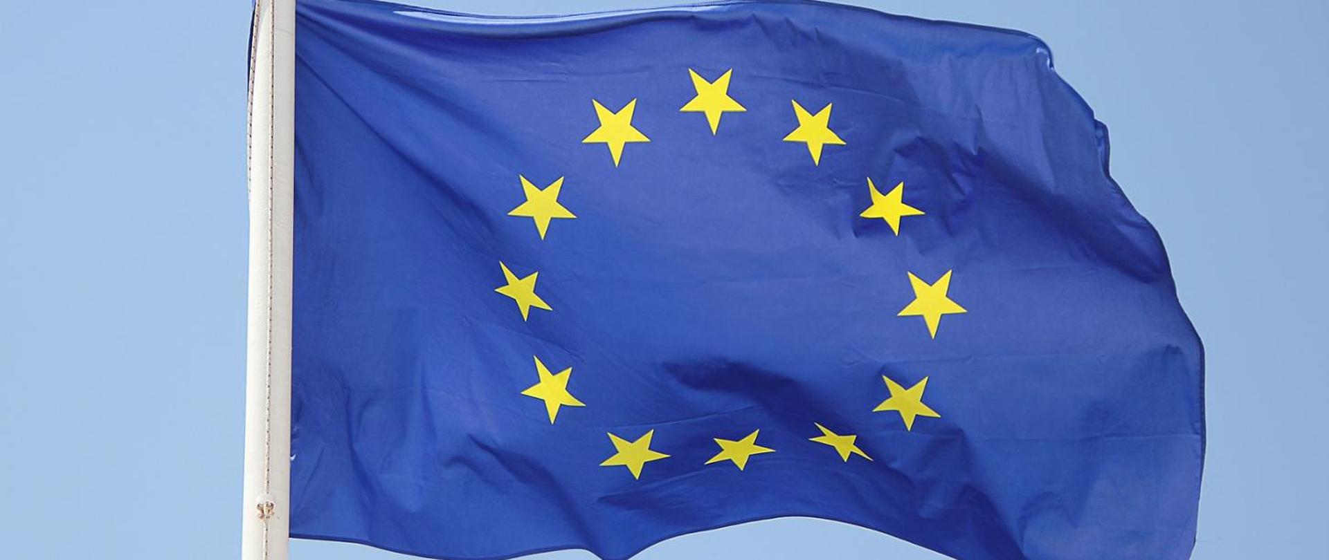 Flaga Unii Europejskiej. Na fladze przedstawiony jest okrąg złożony z dwunastu złotych gwiazd na błękitnym tle.