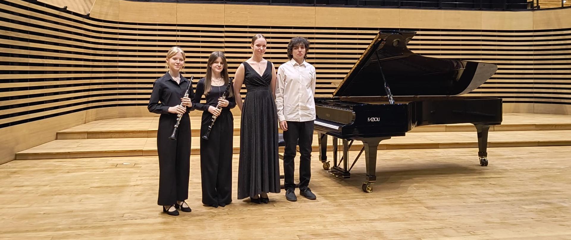 Trzy dziewczyny i jeden chłopak stoją na scenie przy fortepianie, dwie z nich trzymają klarnety.