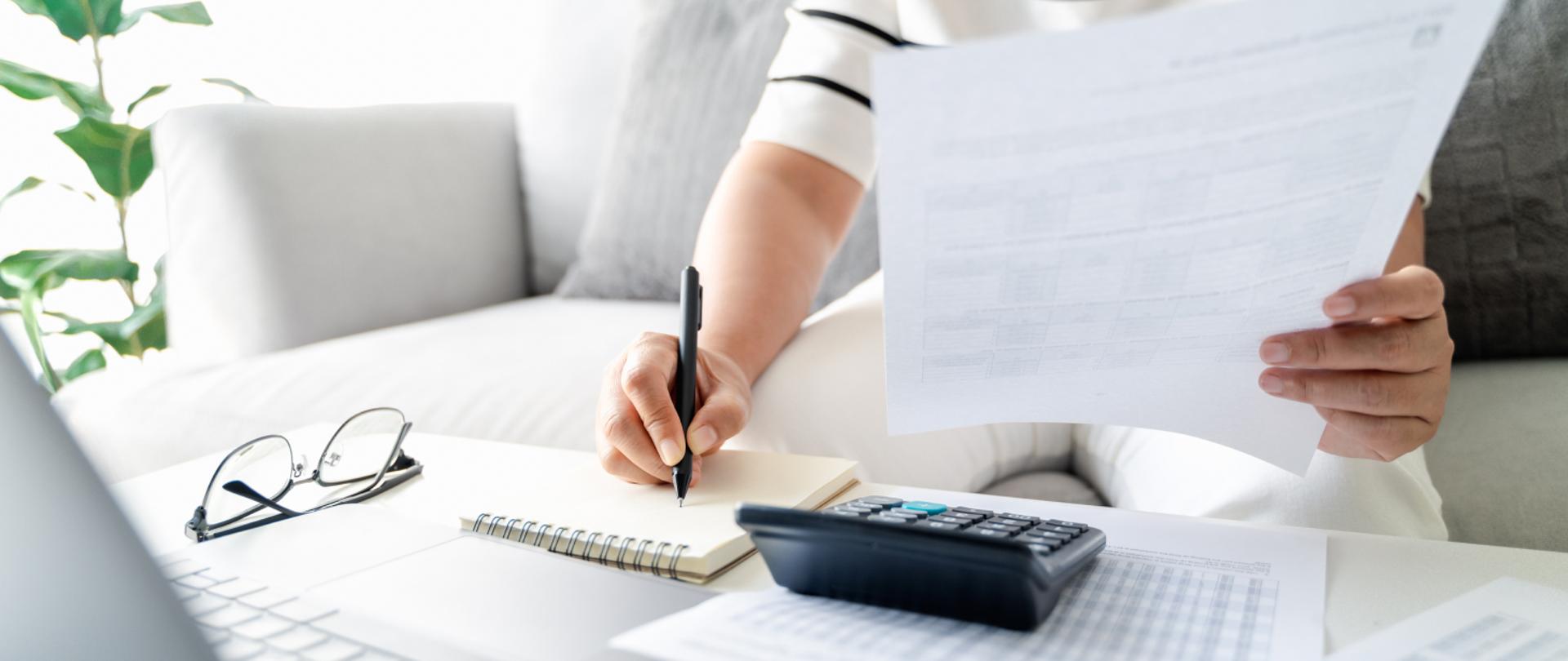Na zdjęciu widać jak ktoś pisze coś na kartce przy biurku. W drugim ręku trzyma dokumenty. Na biurku leżą okulary, kalkulator i inne dokumenty. 