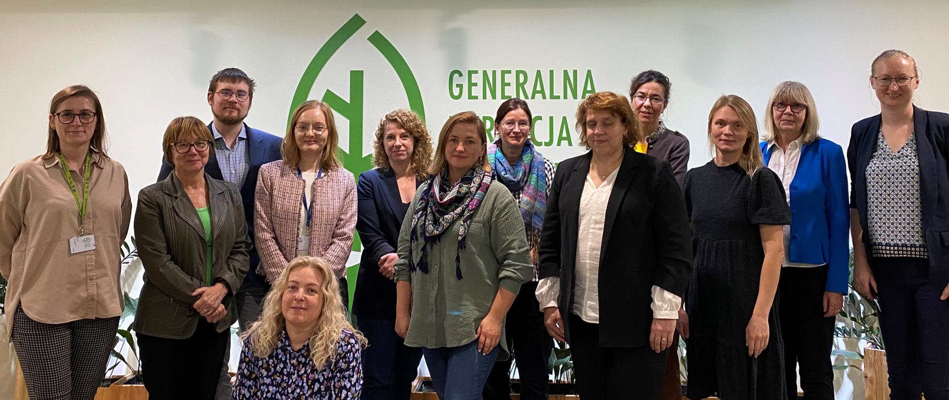 Na tle ściany z logotypem Generalnej Dyrekcji Ochrony Środowiska (zielony liść) stoją kobiety i mężczyźni.