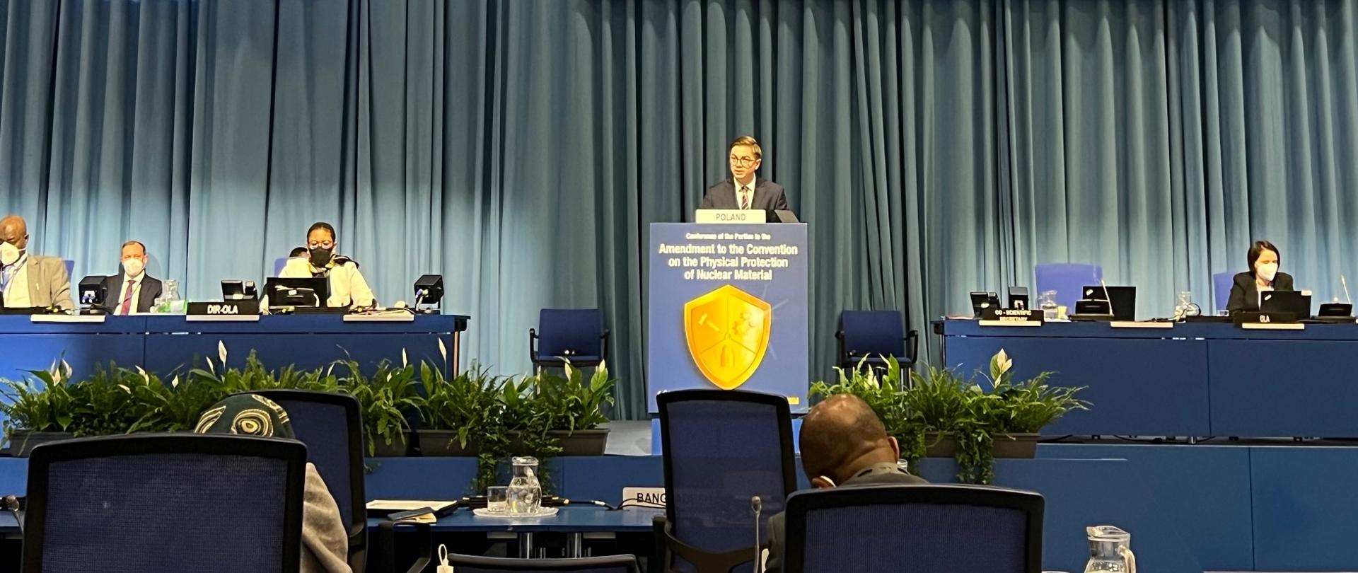 Prezes PAA na konferencji Międzynarodowej Agencji Energii Atomowej w Wiedniu. Dr Łukasz Młynarkiewicz stoi za granatowym pulpitem. Po obu stronach za niebieskimi stołami siedzą uczestnicy konferencji. W tle szara kotara.