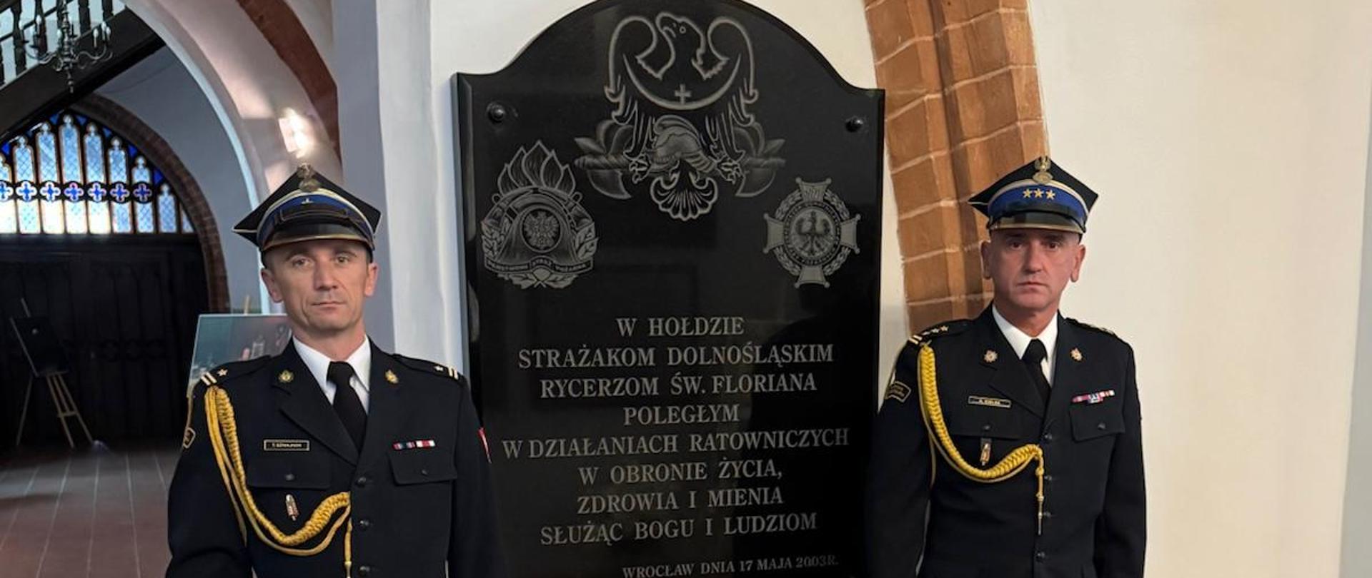 Dwaj strażacy w czarnych mundurach galowych stoją baczność po obu stronach czarnej tablicy pamiątkowej z napisem: „W hołdzie strażakom dolnośląskim rycerzom św. Floriana poległym w działaniach ratowniczych...”. U ich stóp stoi znicz.