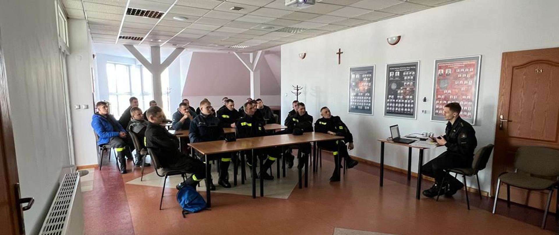 Na zdjęciu widać siedzących w sali ok. 20 osób. Wszyscy siedzą w ławkach wszyscy ubrani są w czarne strażackie stroje koszarowe. Na przeciw nich siedzi przy biurku oficer prowadzący wykłady.
