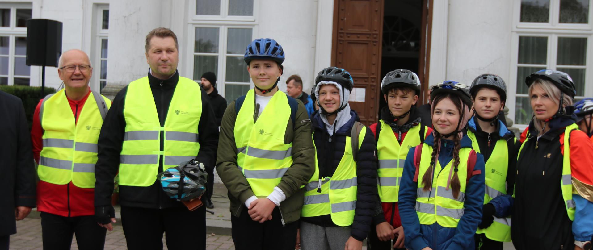 Grupa ludzi w odblaskowych kamizelkach stoi przed białym budynkiem.