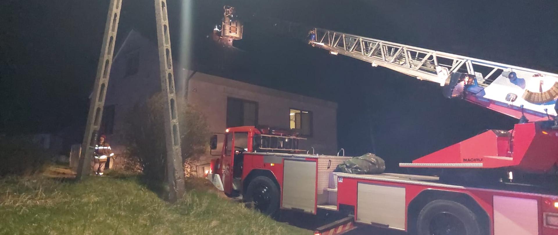 Zdjęcie przedstawia budynek mieszkalny, parterowy. Przed nim znajduje się samochód specjalny z rozłożonym parkiem drabinowym. W jego koszu znajduje się strażak w umundurowaniu specjalnym z czerwonym hełmem na głowie. Strażak pracuje przy obtrącaniu sadzy w przewodzie kominowym. Teren działań oświetlony- zdjęcie zrobione w porze nocnej. 