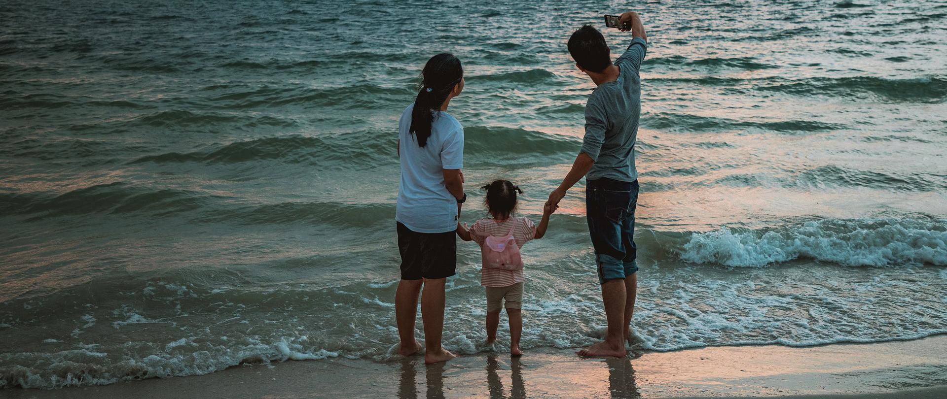 Fotografia rodziny trzyosobowej, w tym małe dziecko, która stoi na brzegu morza. Mężczyzna trzymający za rękę córkę, wykonuje selfie. Po lewej stronie kadru matka dziewczyny, która także pozuje do zdjęcia. W tle otchłań morza oraz w oddali, na niebie, samolot. 