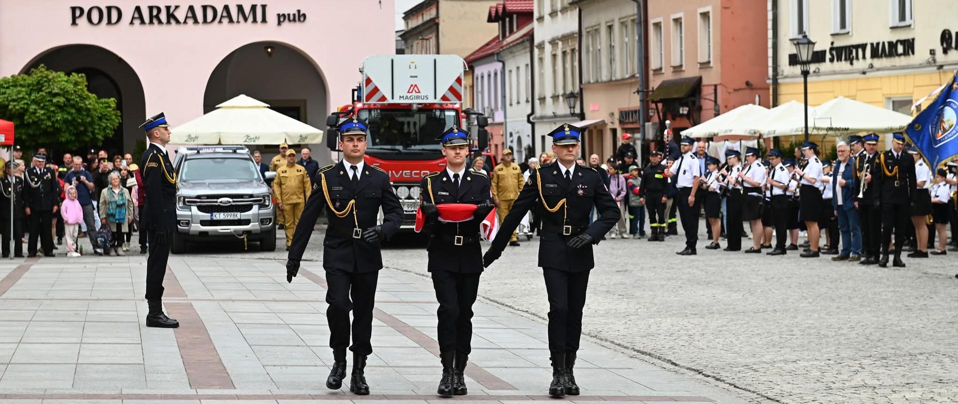Poczet flagowy w postaci trzech strażaków ubranych w granatowe mundury galowe maszeruje po płycie rynku. Środkowy strażak trzyma na rękach bialo-czerwoną flagę. W tle widać kamienice rynku oraz drabinę pożarniczą, obok której stoi orkiestra.