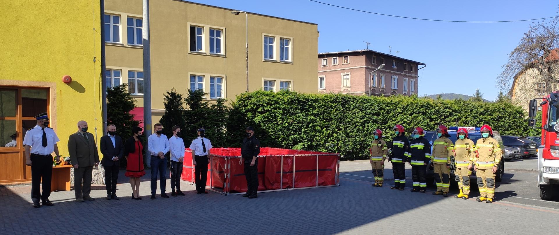 Widok na plac jednostki JRG PSP w Bystrzycy Kłodzkiej. Po lewej stronie w szeregu stoi sześciu strażaków w umundurowaniu specjalnym. Po środku widok na rozłożony basen strażacki koloru czerwonego. Przed nim stoi dowódca JRG w Bystrzycy Kłodzkiej. Po prawe stronie Komendant Powiatowy PSP w Kłodzku w umundurowaniu wyjściowym. Obok w szeregu stoi sześciu zaproszonych gości.