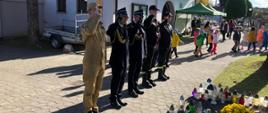Widok z przodu. Funkcjonariusze PSP w szeregu salutujący przed niewidocznym na zdjęciu pamiątkowym krzyżem cmentarnym. W tle kaplica cmentarna. Chodnikiem przechodzą przedszkolaki. Pod krzyżem wiele zniczy oraz żółta chryzantema w donicy. 